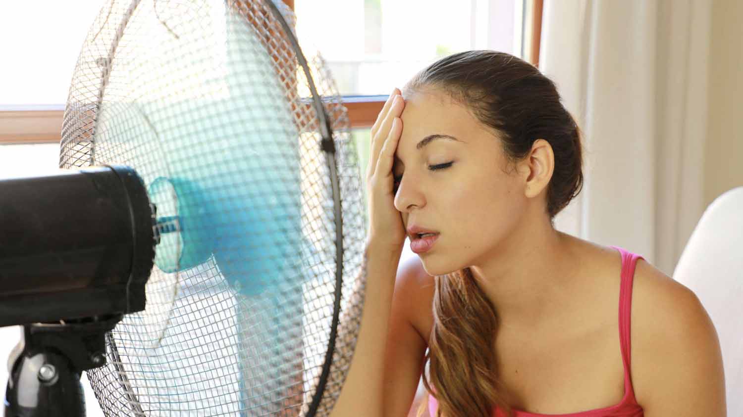 Woman’s thermostat isn’t working right and her house is too hot. She’s cooling off in front of a fan.