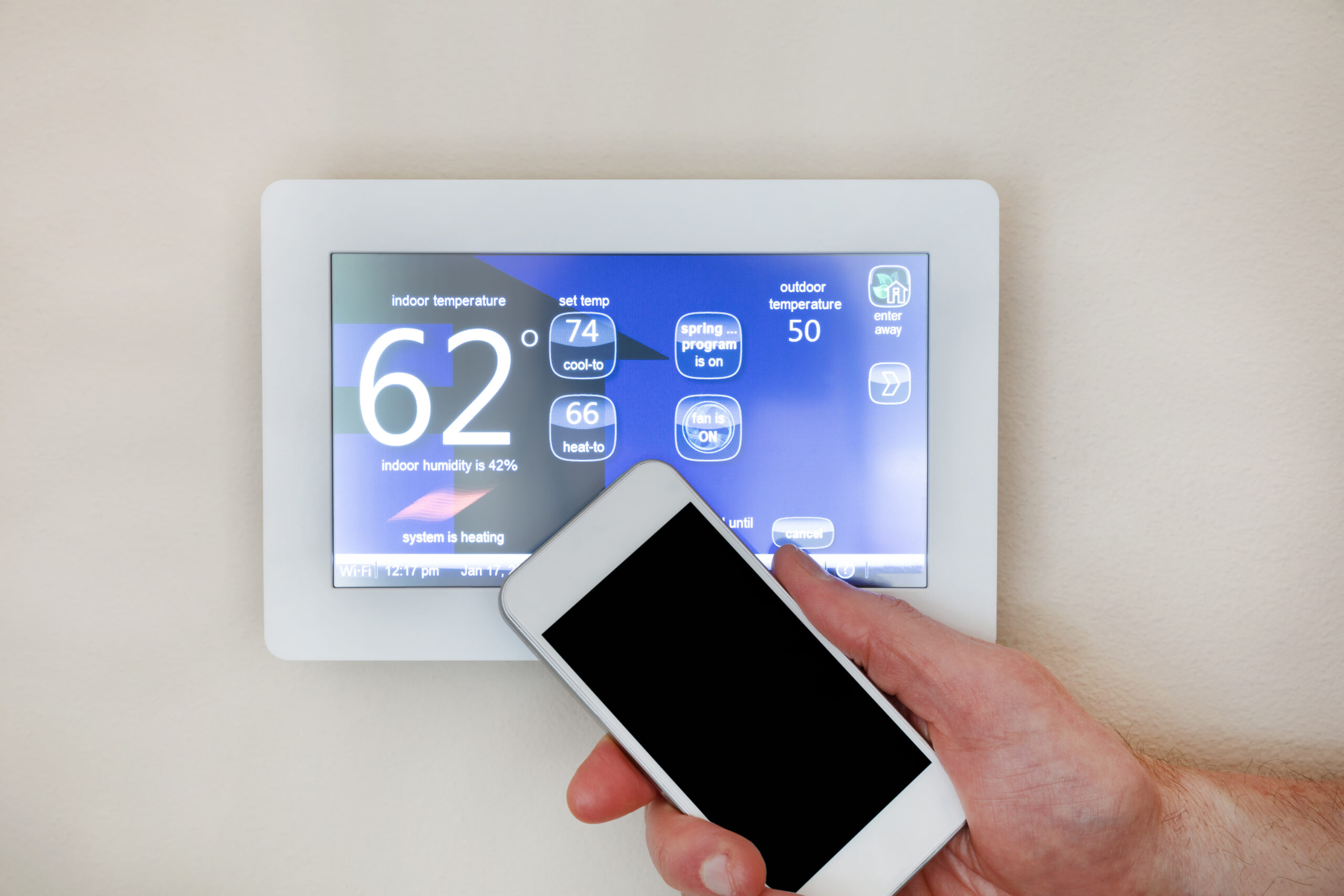 Male hand holding smart phone to operate heating or cooling via digital touch screen programmable thermostat for home