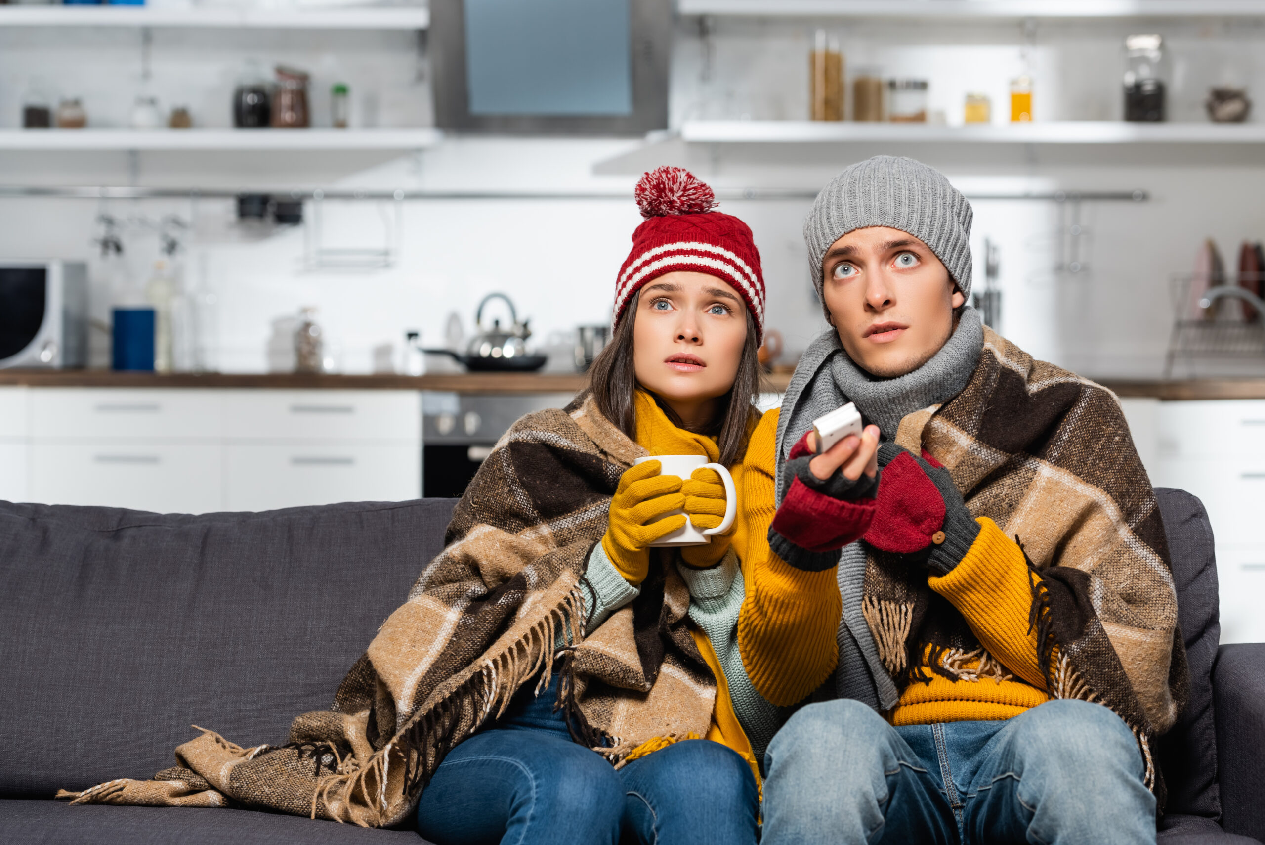 freezing couple wearing hats and gloves, wrapped in plaid blanket, watching tv while sitting in cold house