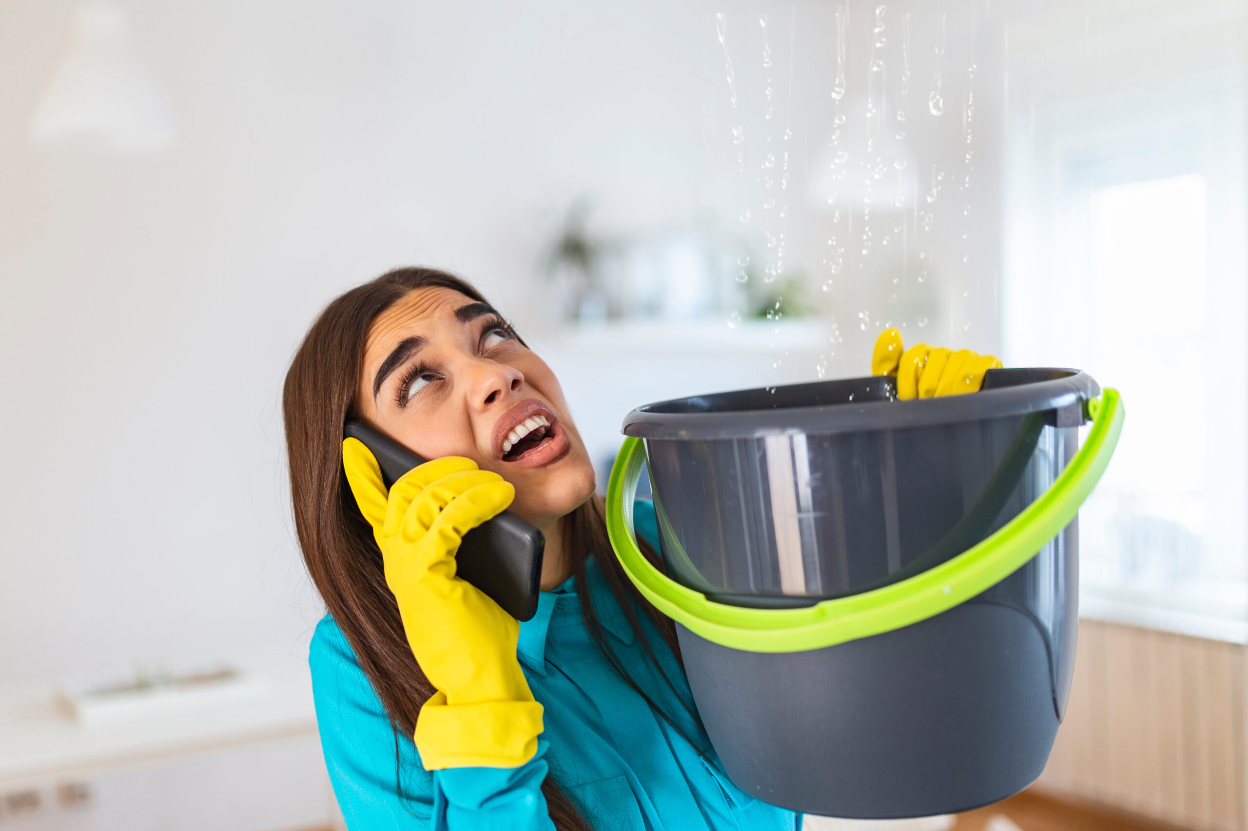 Worried young woman calling plumber while water is leaking from the ceiling that she is catching in a bucket