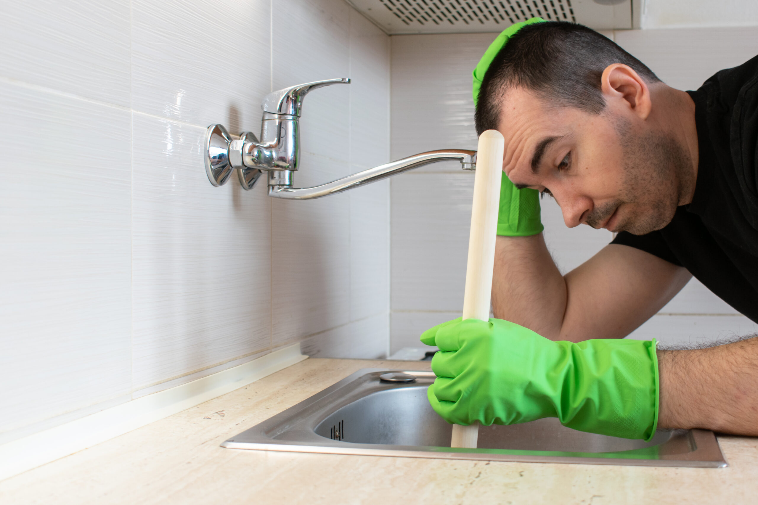 Man wearing green rubber gloves holds a plunger in his clogged kitchen sink