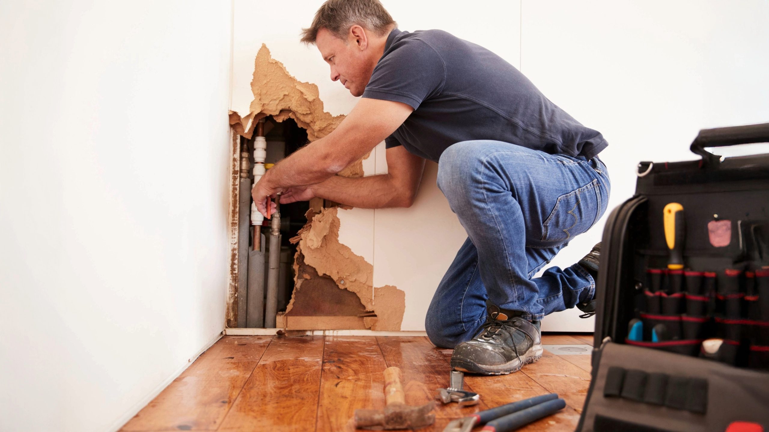 Emergency plumber repairing burst pipe inside wall with visible water damage, tools on floor and open tool bag in foreground